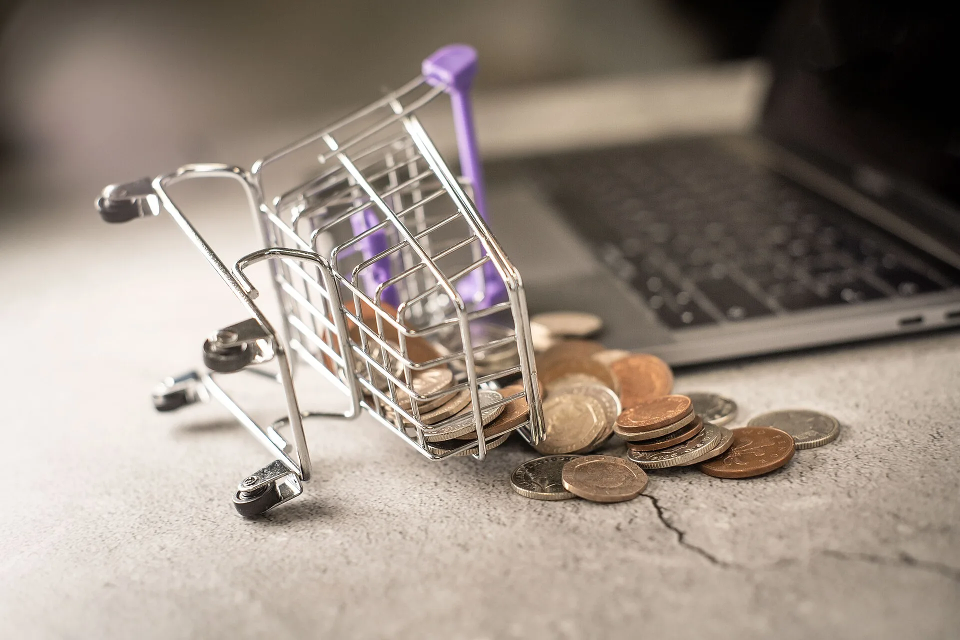 An image of a miniature shopping cart on its side with pennies spilling out of it, representing an abandoned ecommerce cart. The shopping trolley is lying beside an open laptop. This image is free to use under Creative Commons. If you choose to use it, please link back to our website at www.zuko.io. Behavioral trigger email journey ideas that actually fit